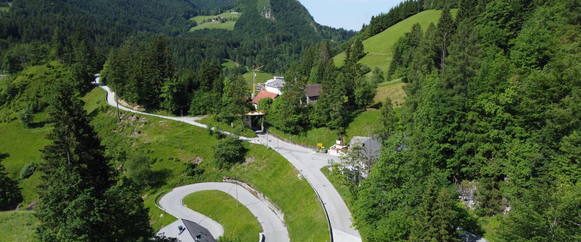 Eine Landschaft mit einem gewundenen Weg, der sich durch grüne Hügel schlängelt, im Hintergrund erheben sich bewaldete Berge, während im Vordergrund ein paar vereinzelte Häuser zu sehen sind