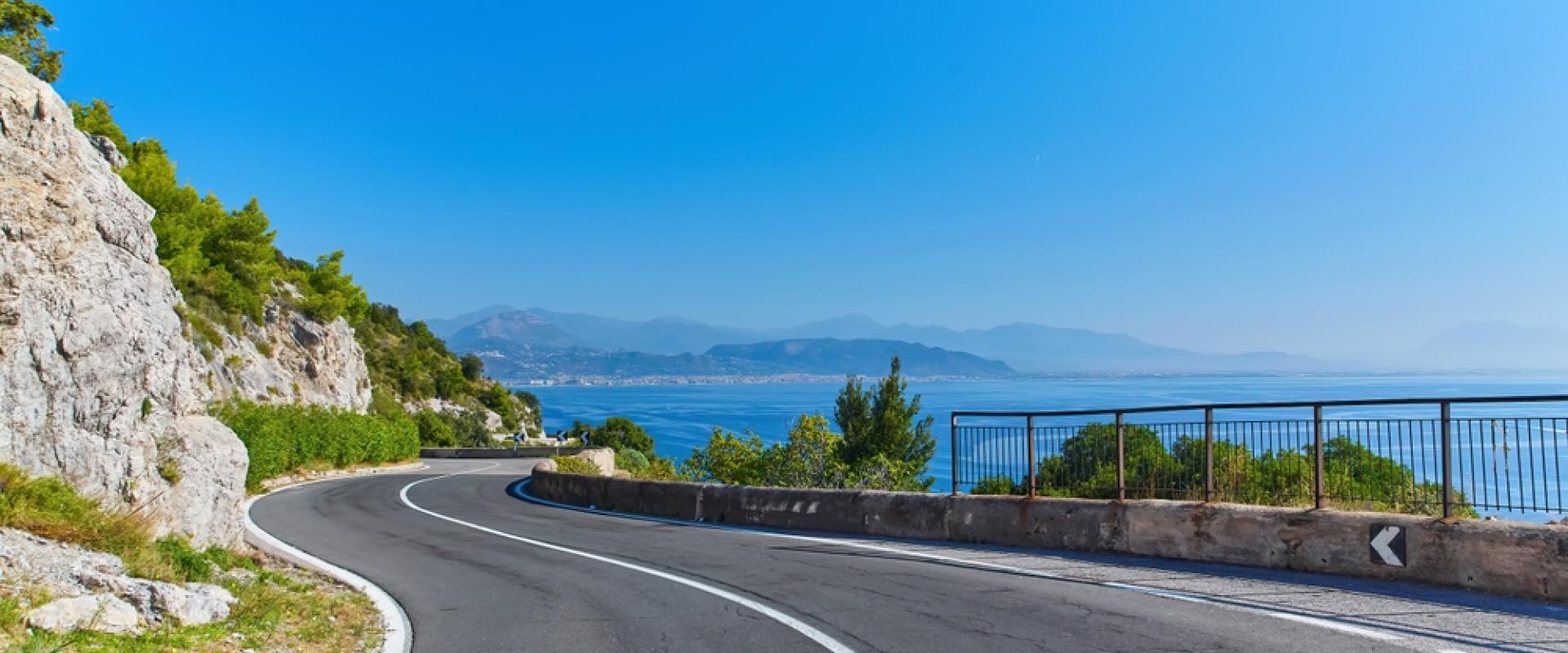 Eine kurvige Küstenstraße führt entlang eines felsigen Hangs, mit Blick auf das blaue Meer und entfernte Berge unter klarem Himmel.