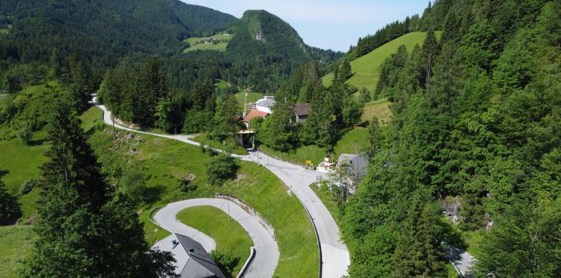 Eine Landschaft mit einem gewundenen Weg, der sich durch grüne Hügel schlängelt, im Hintergrund erheben sich bewaldete Berge, während im Vordergrund ein paar vereinzelte Häuser zu sehen sind