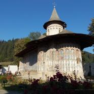 Eine malerische rumänische orthodoxe Klosterkirche mit Spitzdach und aufwendigen Fresken an den Außenwänden, umgeben von einem Garten und Besuchern