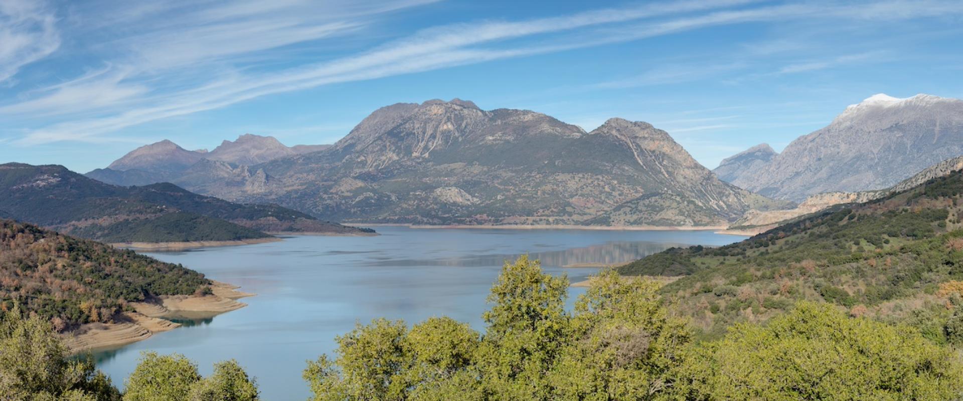 Panoramablick auf den Mornos Stausee in Griechenland in einem Tal, umgeben von Bergen und bewaldeten Hängen unter einem weiten Himmel mit zarten Wolken.