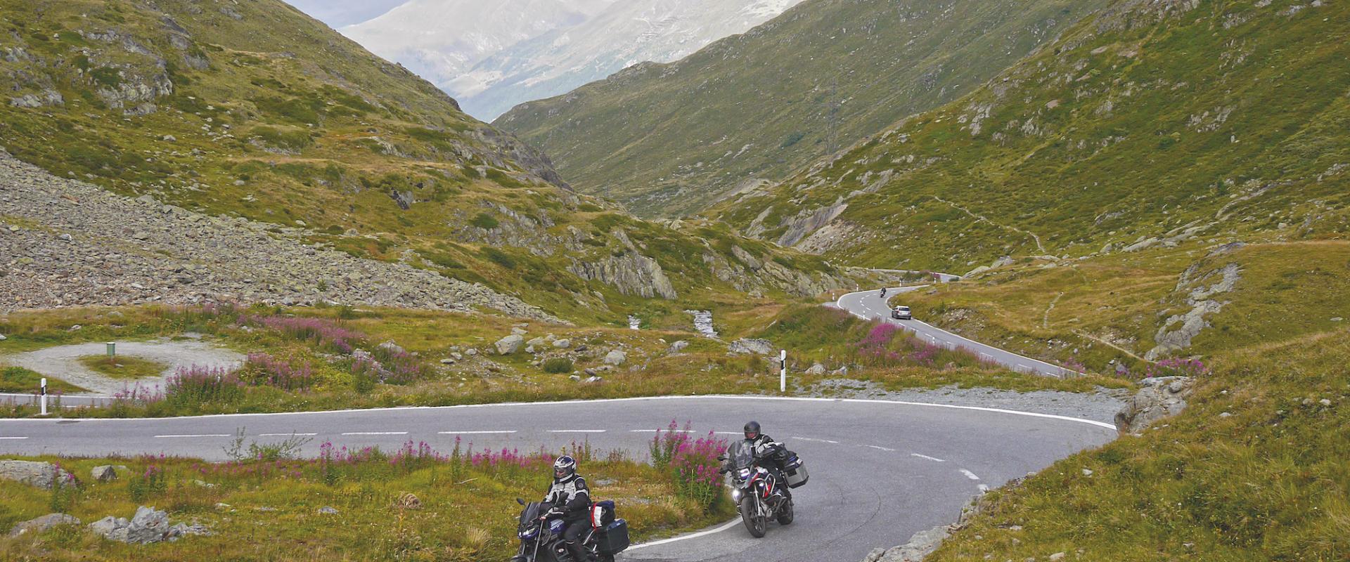 Auf einer kurvenreichen Bergstraße sind zwei Motorradfahrer in einer alpinen Landschaft unterwegs, im Hintergrund erstrecken sich beeindruckende Berge und ein bewölkter Himmel.