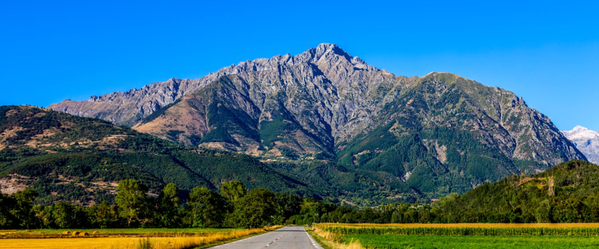 Eine Straße die geradewegs auf ein Bergmassiv zuläuft; rechts und links sind Wiesen und Felder