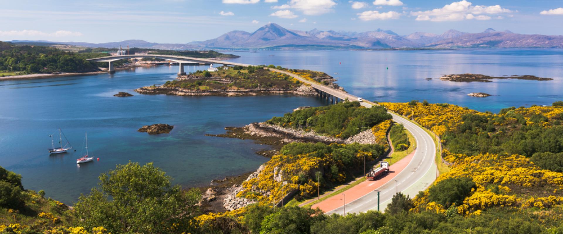 Eine Küstenstraße mit gelben Blumen schlängelt sich durch grüne Hügel, vorbei an Segelbooten und einer Brücke mit Bergen im Hintergrund.