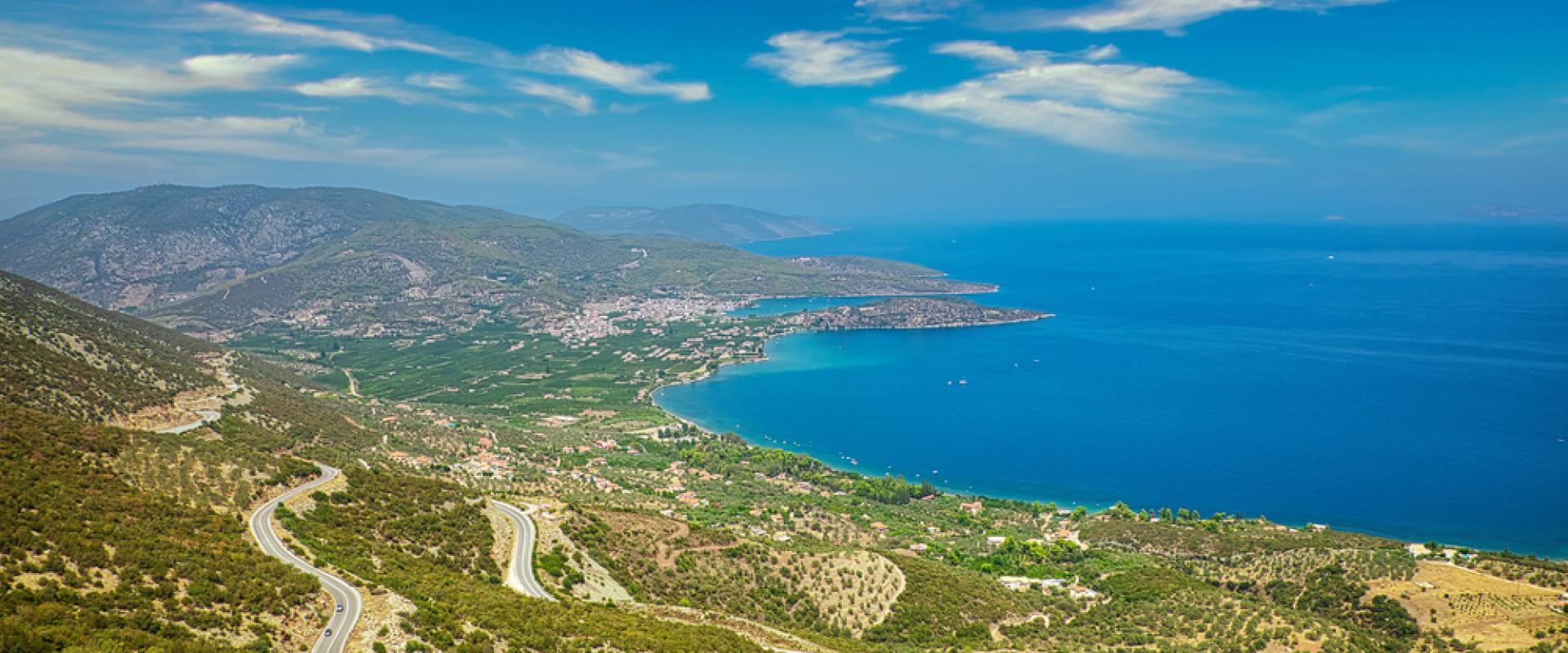 Ein kurviger Straßenverlauf führt durch eine hügelige Landschaft mit Panoramablick auf die griechische Küste auf Peloponessos. Man sieht eine Küstenlinie und das blaue Meer unter einem klaren Himmel.