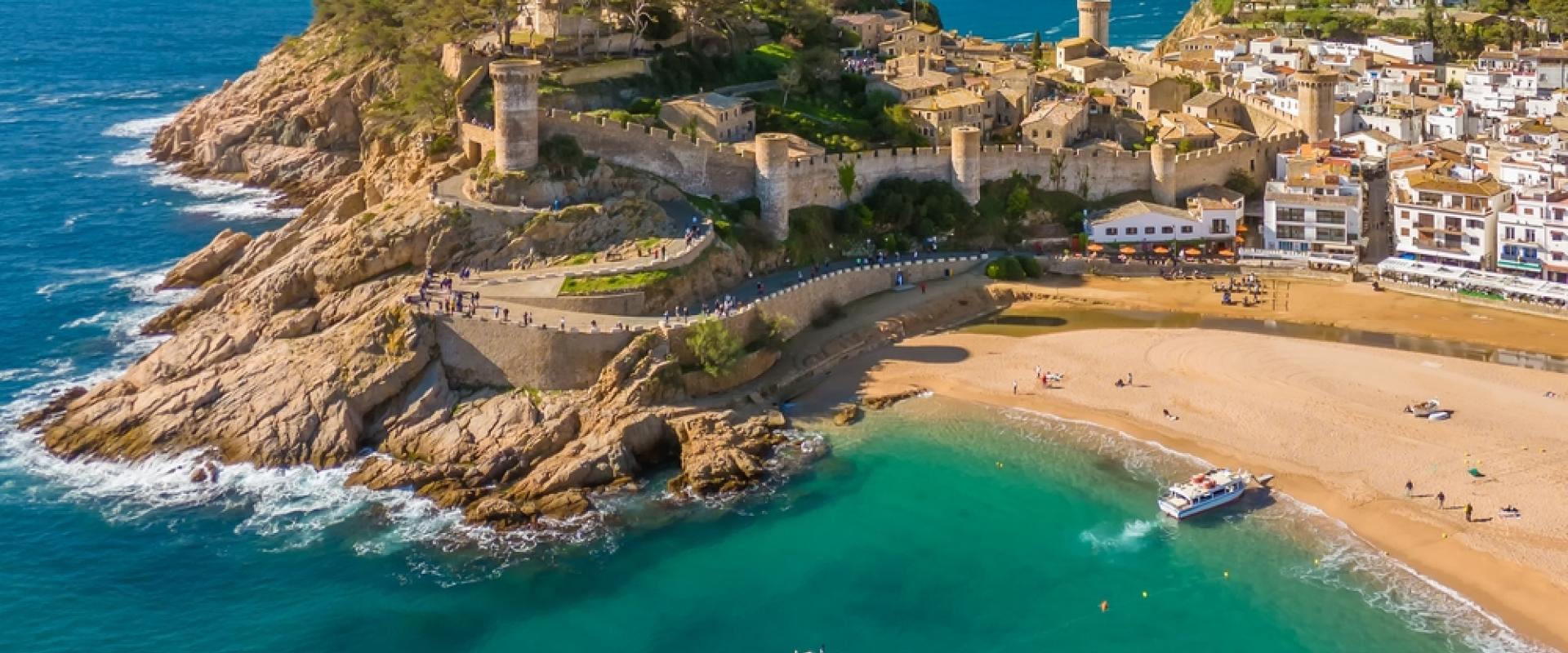 Luftaufnahme einer mittelalterlichen Burg auf einem Hügel am Meer, mit einem Sandstrand und Booten im klaren, türkisfarbenen Wasser.