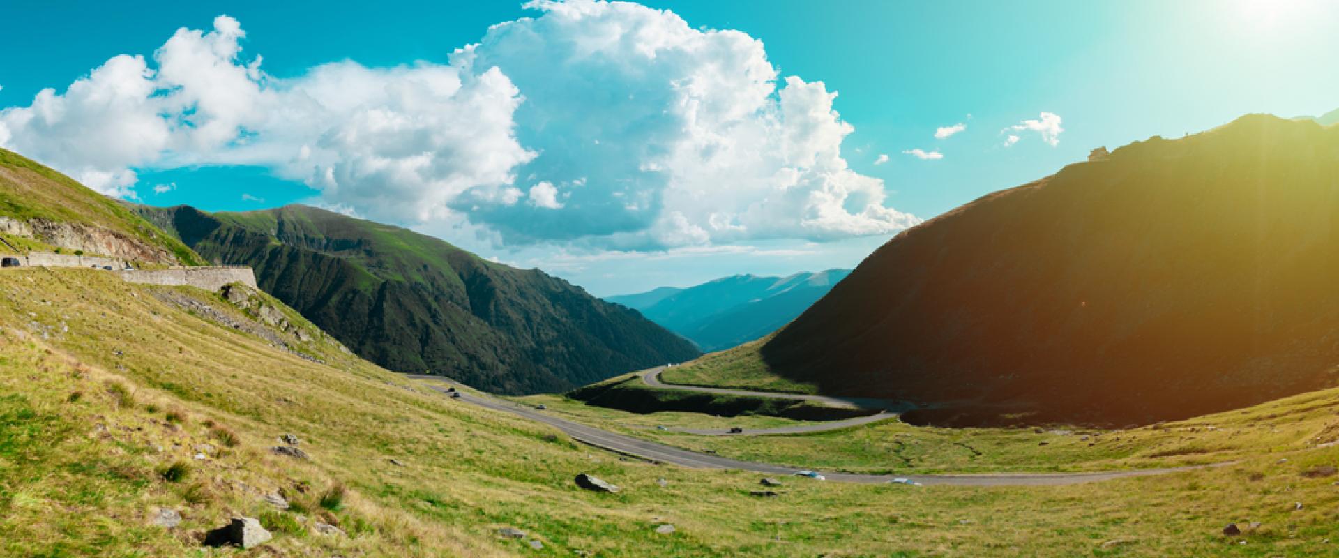 Weitläufige Gebirgslandschaft mit grünen Hügeln und kurviger Straße. Sonnenschein fällt von rechts, große Wolken über den Bergen am Horizont.