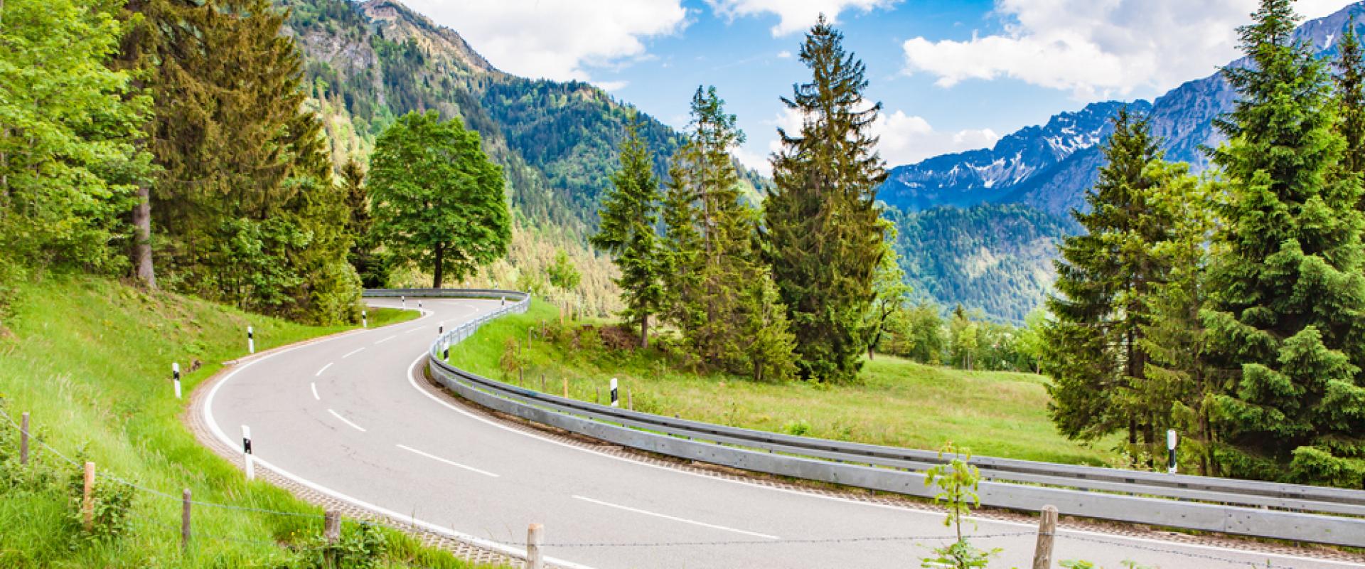 Eine geschwungene Bergstraße führt durch grüne Wiesen und Wälder, im Hintergrund erheben sich schneebedeckte Berge unter blauem Himmel.