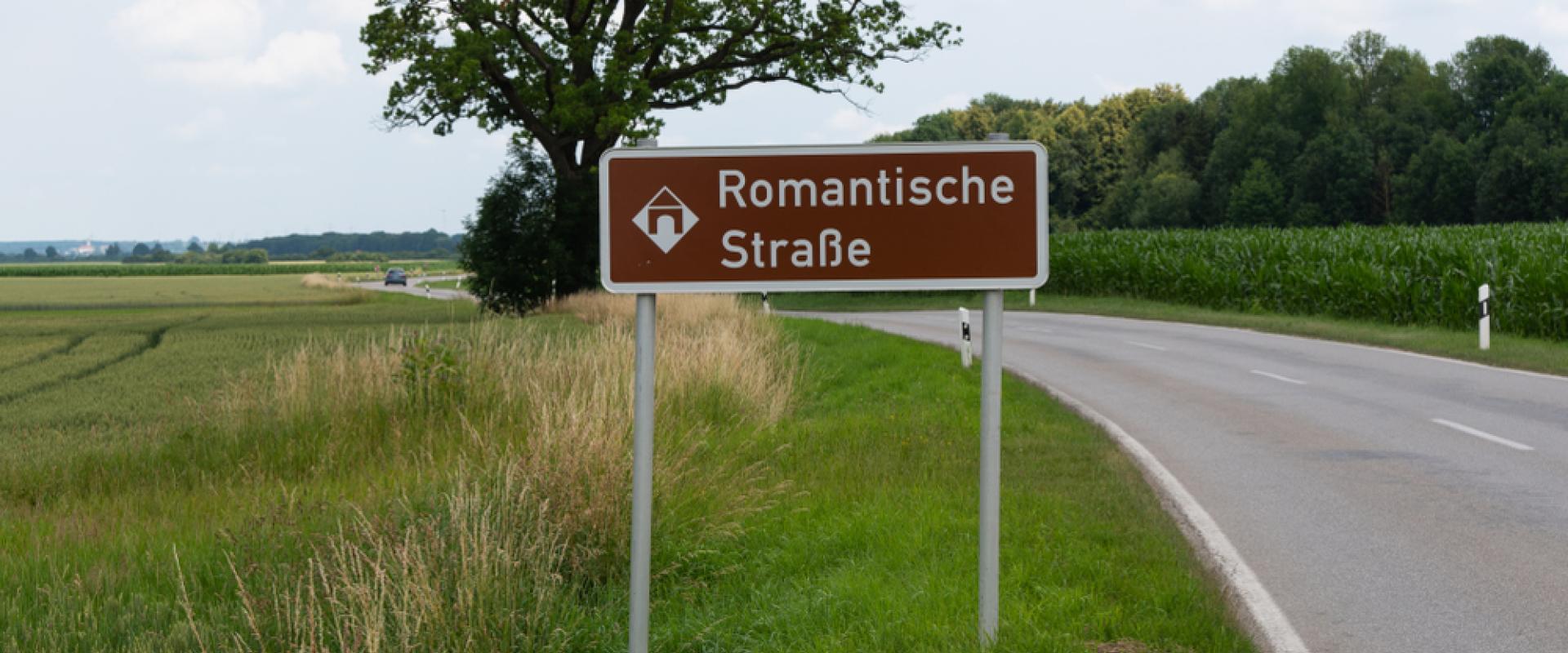 Ein braunes Straßenschild mit der Aufschrift „Romantische Straße“ steht neben einer Landstraße, umgeben von Feldern und Bäumen.