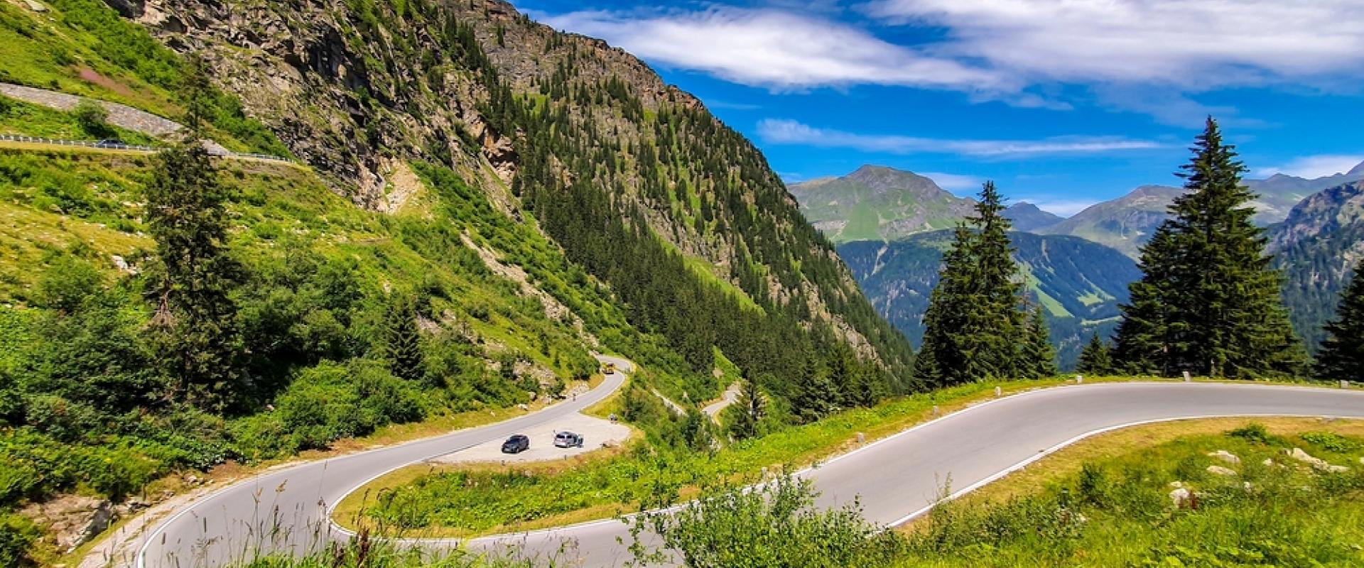 Eine kurvenreiche Bergstraße windet sich durch eine grüne, felsige Alpenlandschaft. Zwei Autos fahren auf der Straße, umgeben von Tannen und Bergen unter blauem Himmel.