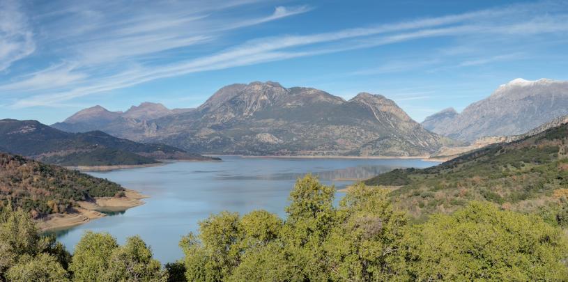 Panoramablick auf den Mornos Stausee in Griechenland in einem Tal, umgeben von Bergen und bewaldeten Hängen unter einem weiten Himmel mit zarten Wolken.