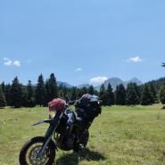 Ein beladenes Touring-Motorrad mit einem rot kariertem Schal am Lenker steht bei einer  Motorradtour durch Griechenland auf einer Wiese in einer bergigen Landschaft mit Nadelbäumen im Hintergrund bei klarem Himmel 