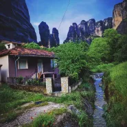 Ein traditionelles Haus an einem kleinen Fluss mit den hohen Meteora-Felsen im Hintergrund, umgeben von grünen Bäumen und Gräsern