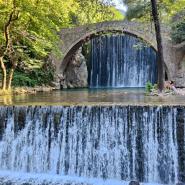 Ein alter Steinbogenbrücke mit einem Wasserfall im Vordergrund, umgeben von grünen Bäumen