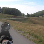 Motorradperspektive einer kurvigen Landstraße im Apuseni Gebirge, die sich durch eine hügelige Landschaft mit vereinzelten Bäumen, wiesen und Feldern zieht